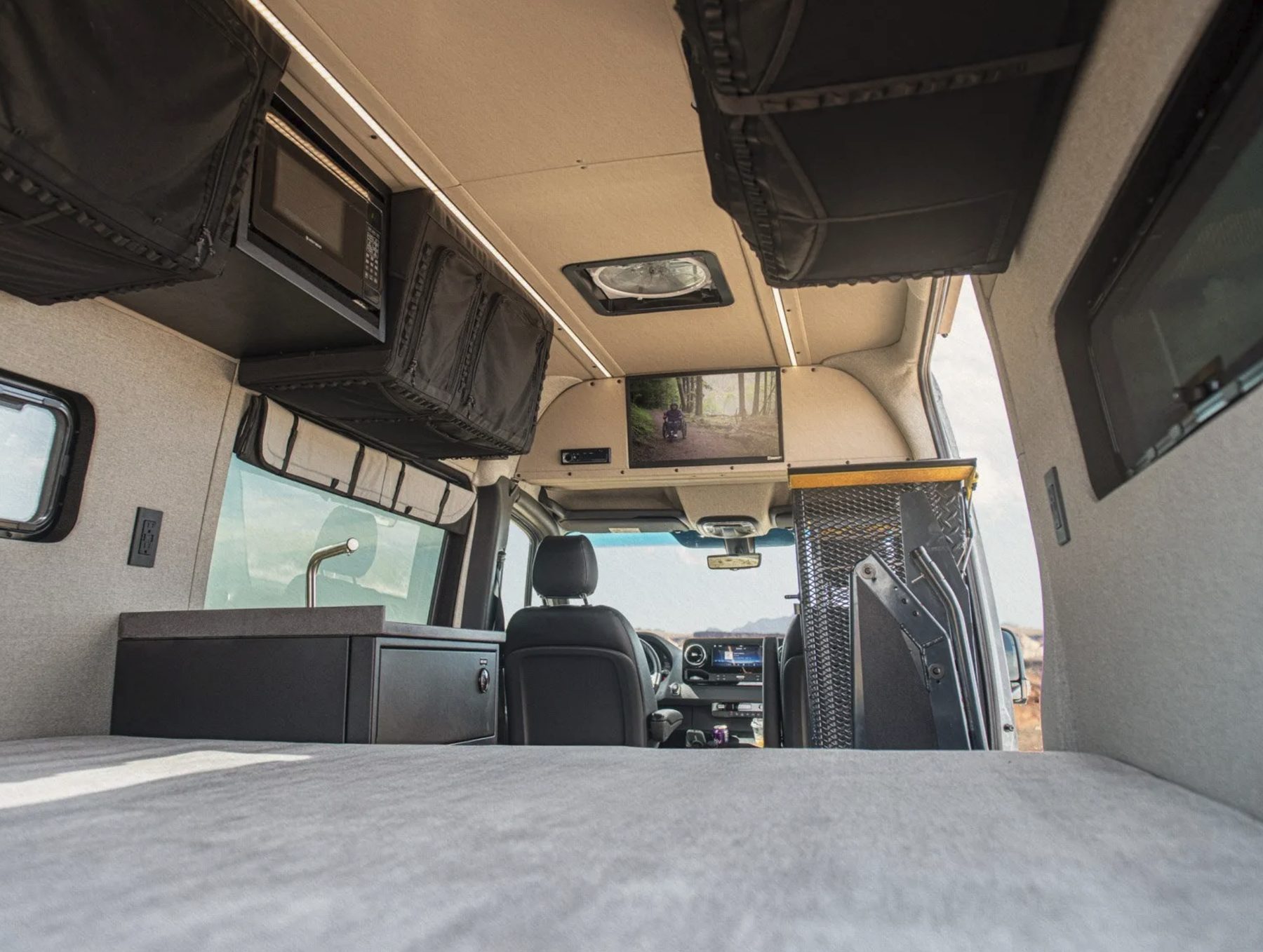 The inside of a mobility camper van photographed from the bed looking forward: overhead storage and a microwave above a galley counter, a wheelchair lift folded against the open side door, the cab seats beyond, and desert visible through the windscreen.