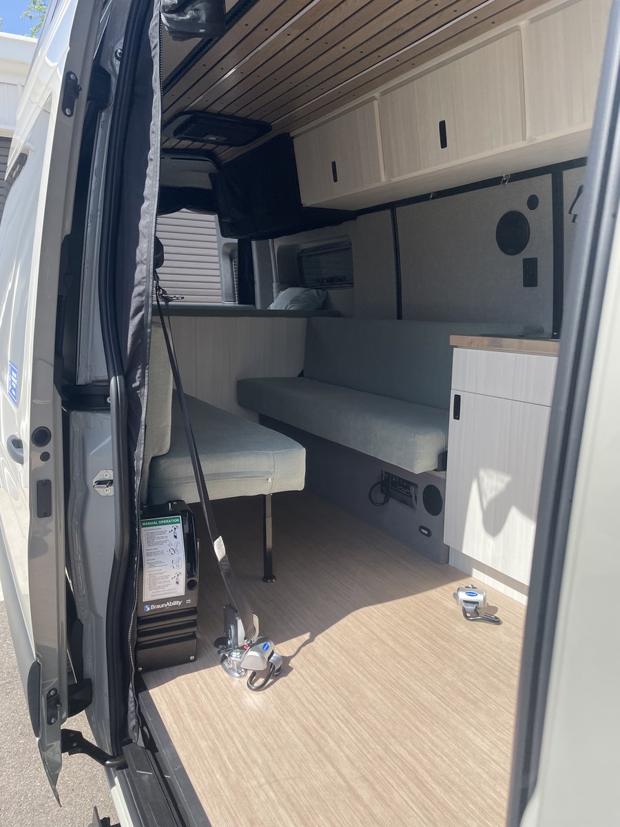 View through the open side door: a long upholstered bench seat with a fold-out extension, a galley cabinet with a timber worktop, a made bed on a raised platform behind, overhead cabinets, and the stowed BraunAbility lift in the foreground beside the door.