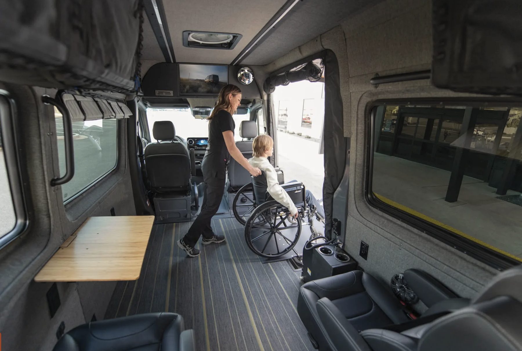 Inside a fully custom mobility van: a woman guides a wheelchair user in through the open side door onto carpeted flooring, past a fold-down wooden table, captain&rsquo;s chairs, overhead curtains and a mounted screen. Seating and open floor are arranged around the wheelchair position.