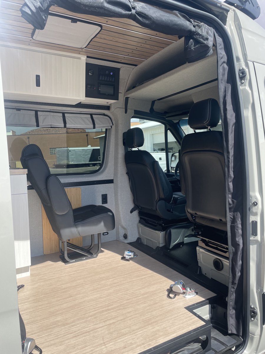 Inside the van looking toward the cab: a black leather captain&rsquo;s chair on a swivel pedestal, the driver and passenger seats beyond it, overhead cabinets, a wall-mounted electrical control panel with switches and a monitor, and a wide clear wood floor with flush securement anchors.