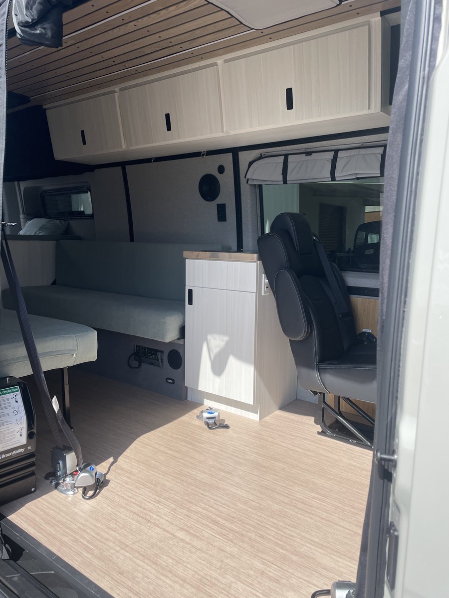 The side doorway of the van with a BraunAbility lift folded upright and stowed against the left door frame, its manual-operation instruction placard facing out. Inside: a bench seat, a galley cabinet with a timber top, an upper bed platform, and two recessed securement anchors in the wood floor.