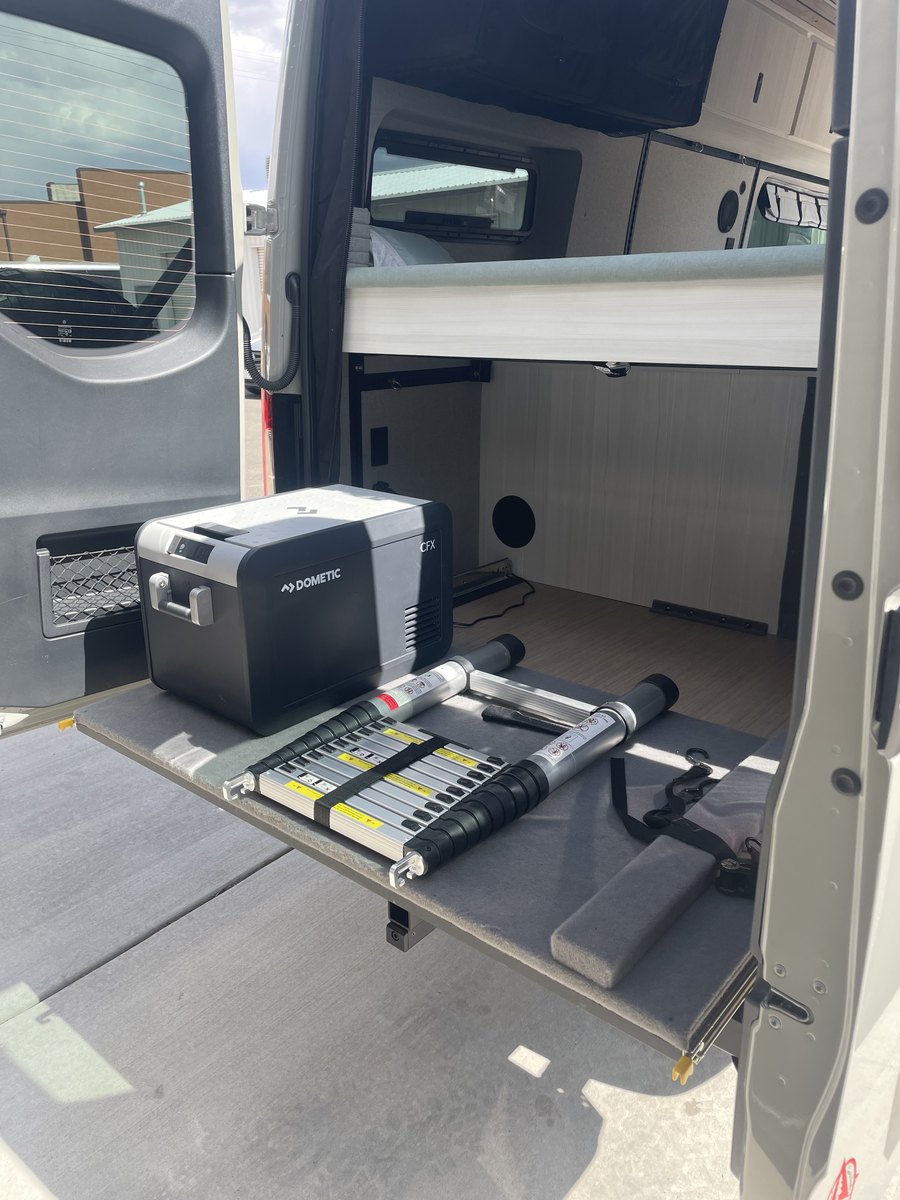 The rear doors open onto a slide-out tray at waist height carrying a Dometic portable fridge and a folded telescopic ladder, with the bed platform above it and an open storage bay beneath.