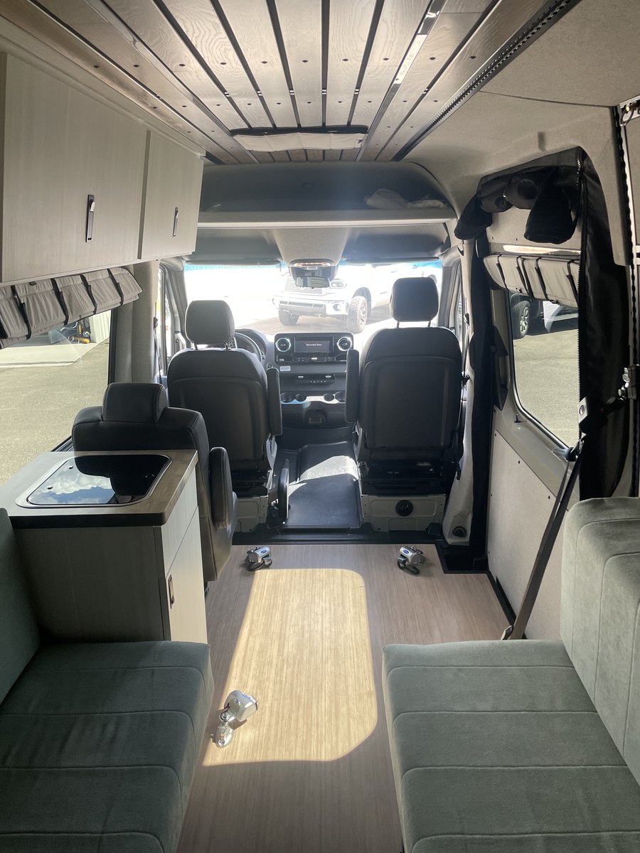 Looking forward from the rear of the van: two facing upholstered bench seats with a clear wood-floor aisle between them, a galley counter with an inset sink on the left, the Mercedes-Benz cab and swivelling front seats ahead, and a slatted timber ceiling above. Four recessed floor anchors sit in the open aisle.