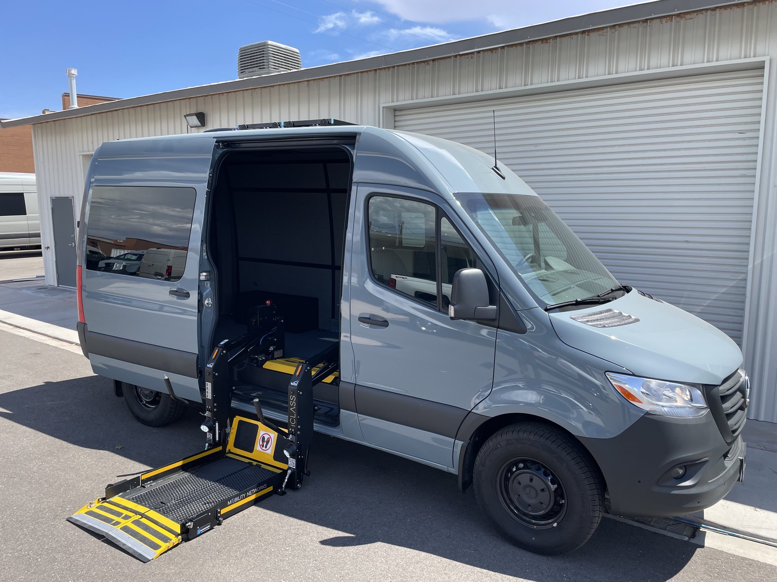 A grey Mercedes-Benz Sprinter parked outside the shop with its side door open and a platform lift fully deployed to the ground, the yellow roll-stop folded flat so a chair can roll straight on.