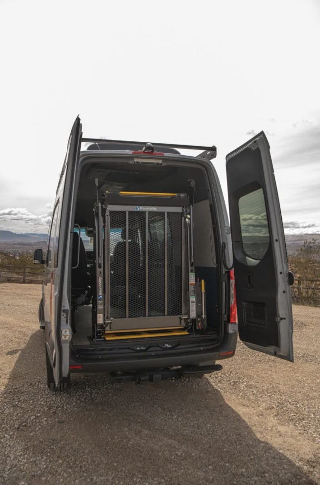 The same Sprinter with its rear doors open and the BraunAbility lift folded upright and stowed flat against the inside of the doorway, leaving the van&rsquo;s floor and rear opening clear.