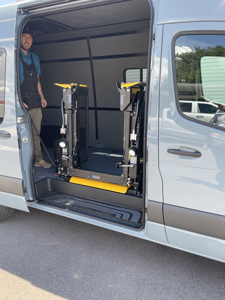 A Guide Mobility technician standing inside the empty Sprinter beside the newly fitted lift, which is folded upright in the side doorway, the pendant control cable hanging from his hand.