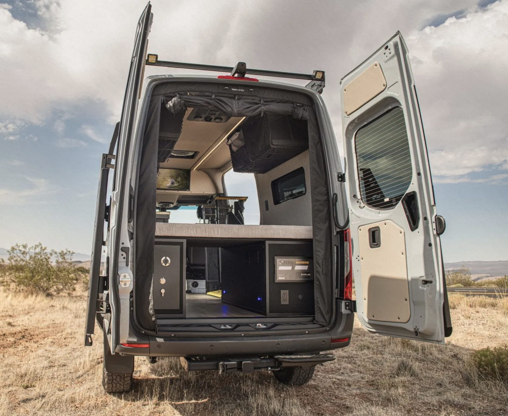 A mobility camper van in open desert with both rear doors swung wide, showing a raised bed platform above a galley bay with a power station and storage, a wheelchair lift folded inside, and overhead cabinets and curtains.