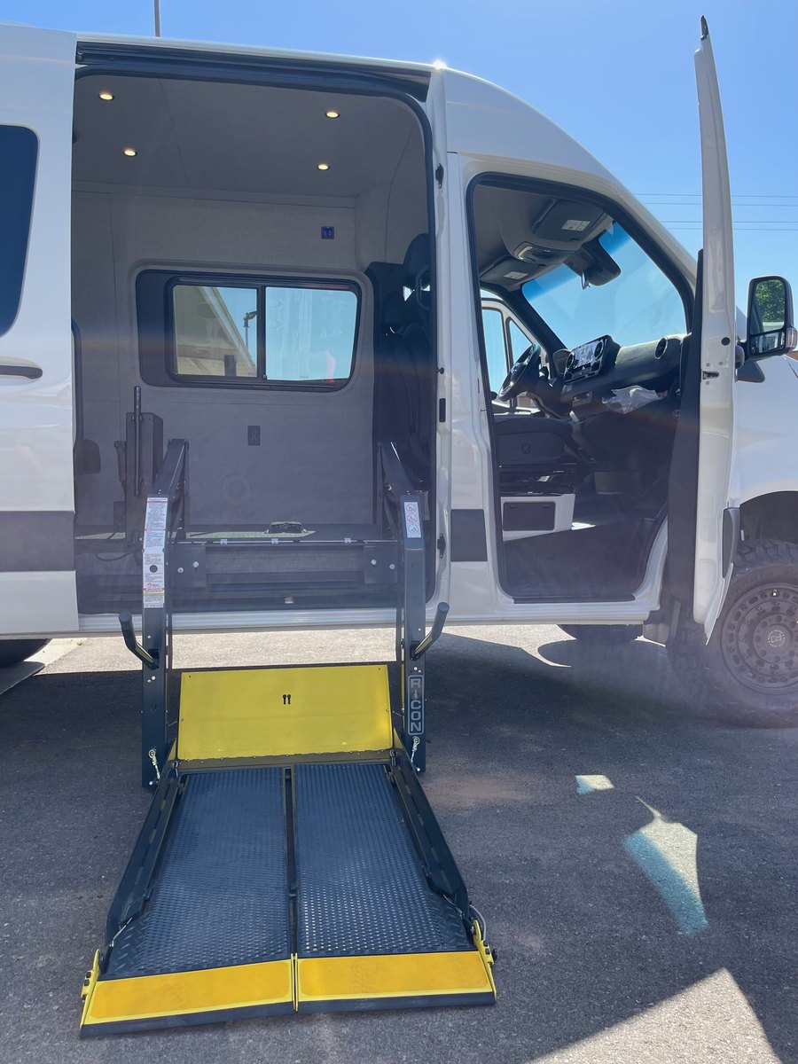 The same RICON lift deployed flat on the asphalt beside the white Sprinter, its yellow roll-stop folded down at the outer edge and the cab door standing open.