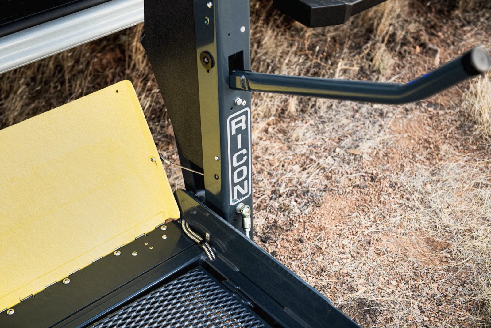 Close view of a RICON lift platform at ground level, showing the yellow roll-stop plate folded flat, the mesh deck and the grab handle &mdash; the hardware a technician inspects at every service.
