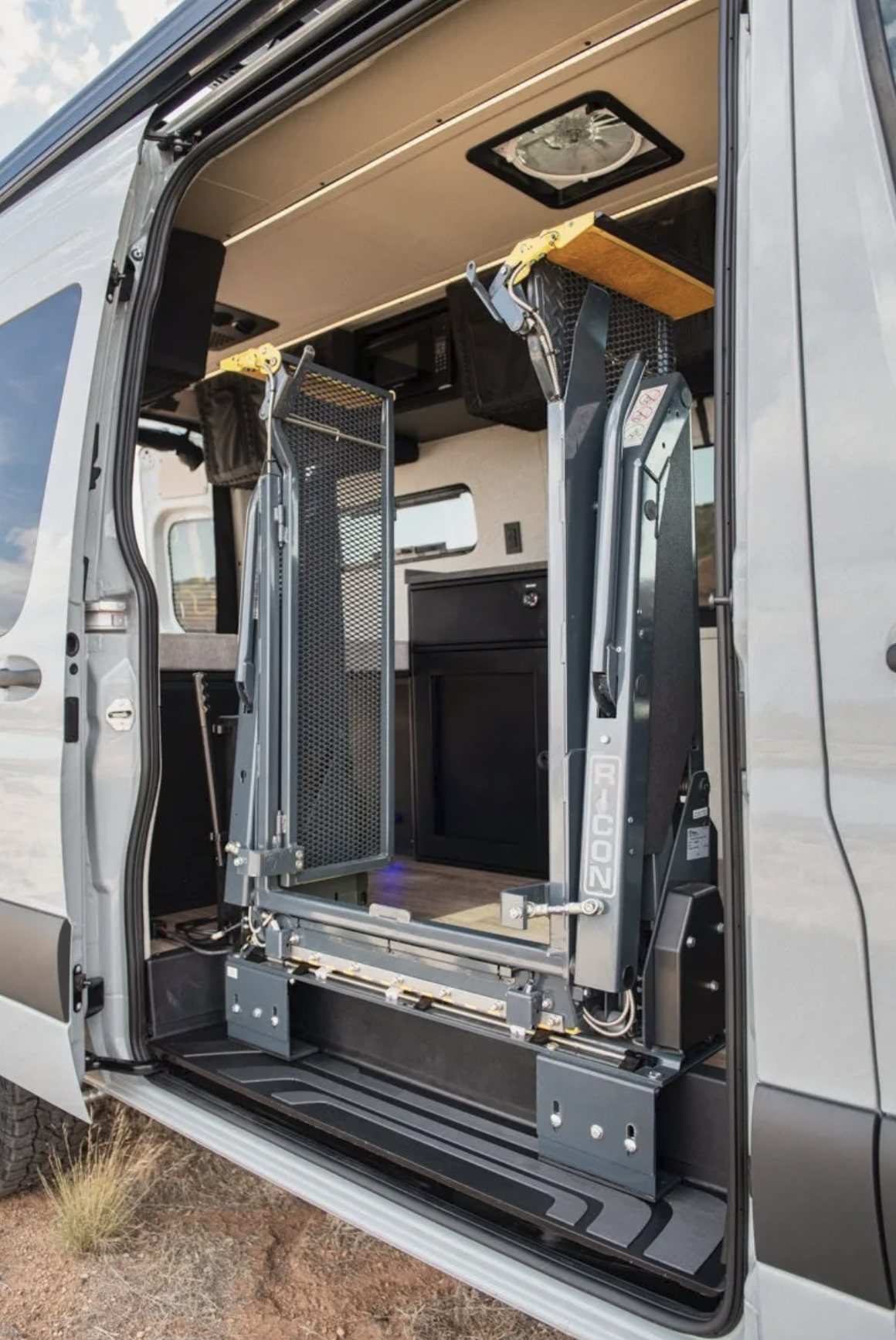 A RICON side-entry lift in one of the mobility camper vans, folded upright in its stowed position against the open side door, with the camper&rsquo;s galley and cabinetry visible beyond it.