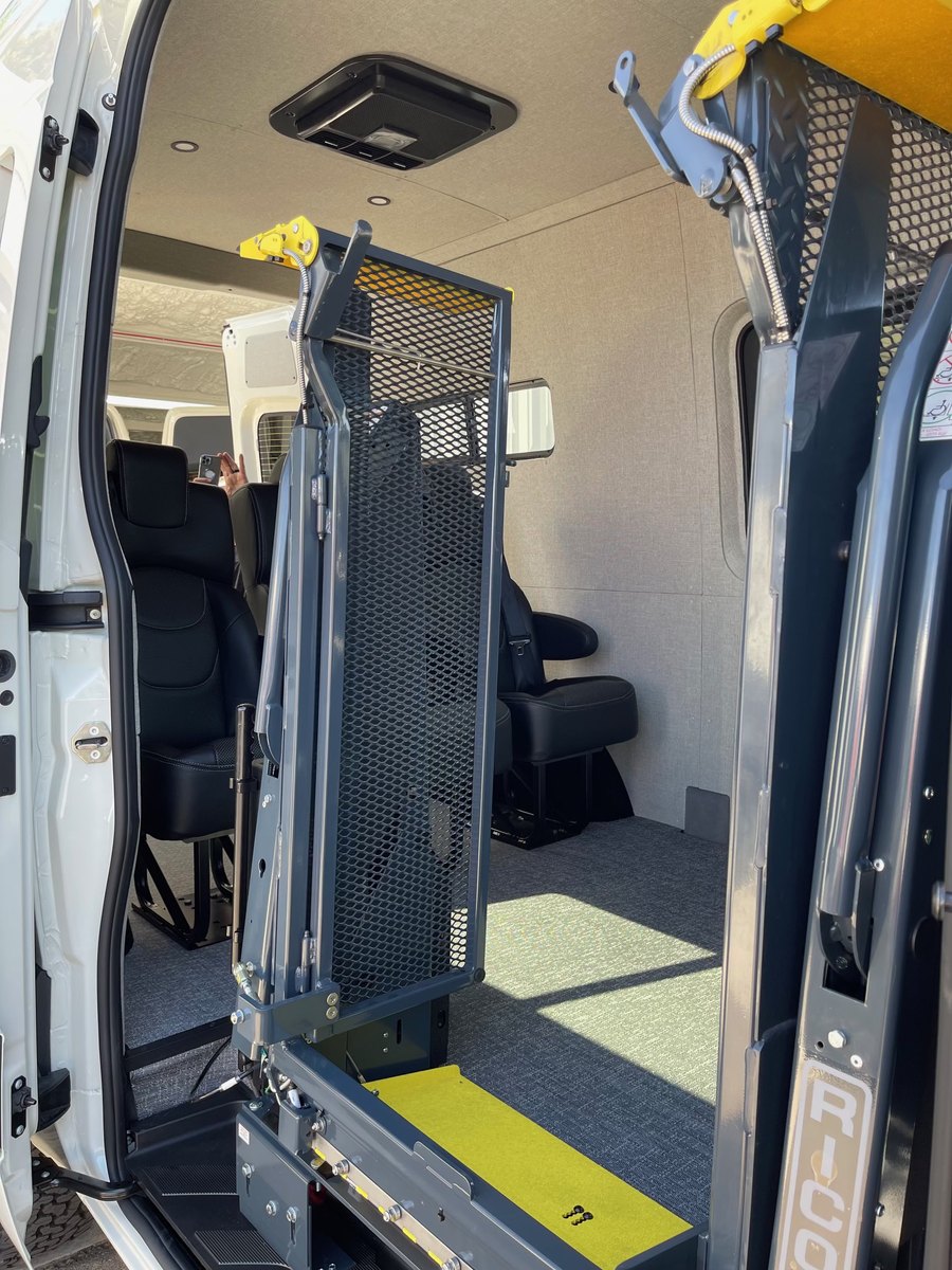 The RICON platform lift folded upright and stowed inside the side doorway of the transport van, its mesh platform vertical and yellow roll-stop at the top, with passenger seats and the cab visible beyond.