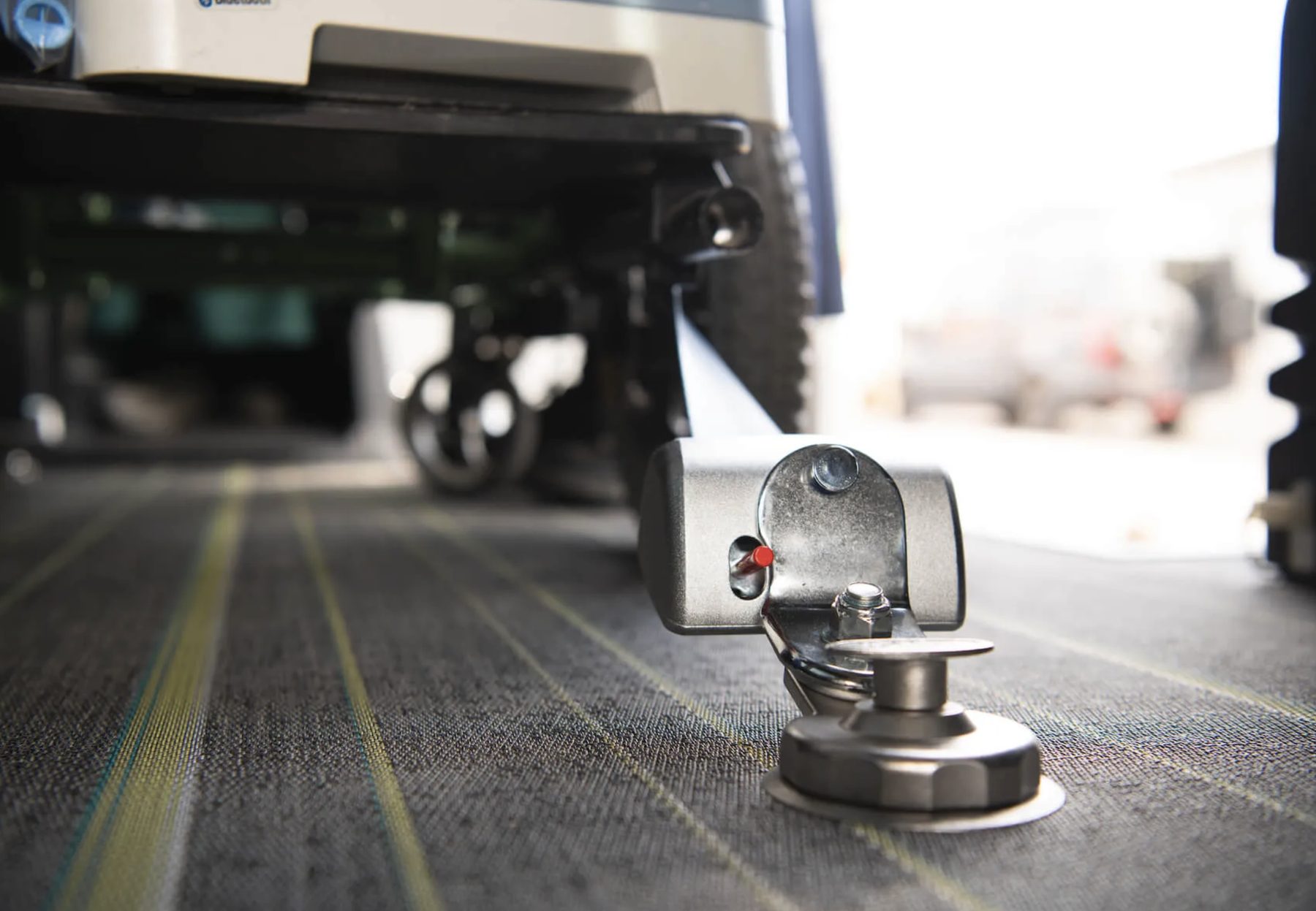A retractable wheelchair securement anchor locked into a recessed floor track, photographed at floor level with the wheels of a secured wheelchair behind it.
