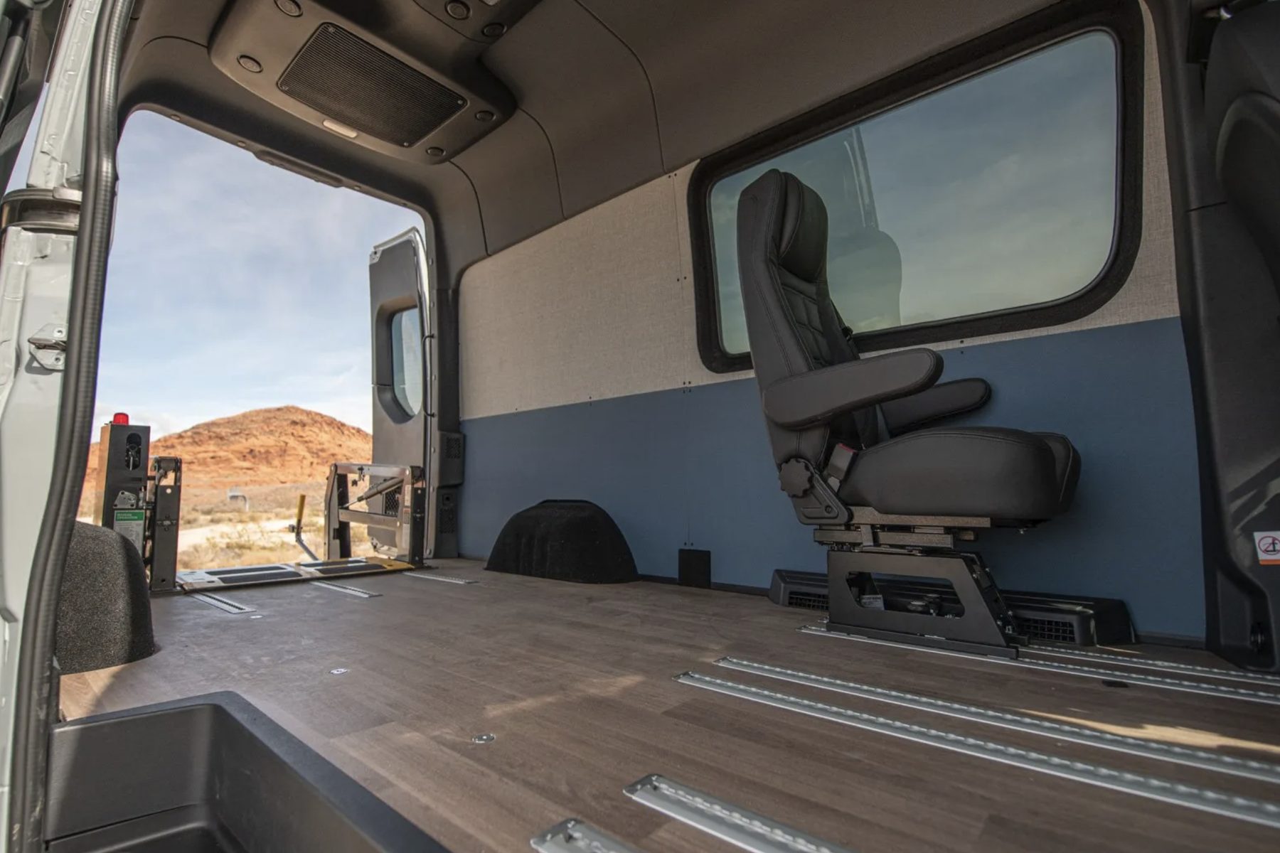 Inside the Sprinter looking toward the rear: a wide wood-grain floor with four recessed L-track securement rails running its length, a black captain&rsquo;s chair on a swivel base against the cream-and-blue upholstered wall, and the side door open onto red rock country with the wheelchair lift visible at the back.