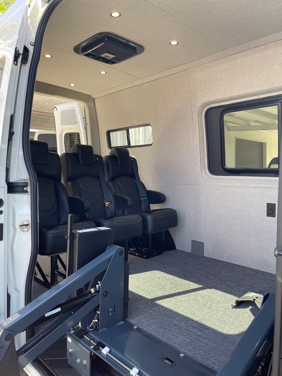 Inside a transport Sprinter: two rows of black forward-facing passenger seats along the left side, a wide open securement bay with floor anchors on the right, grey carpet-textured flooring, and the RICON lift arm at the open side door.