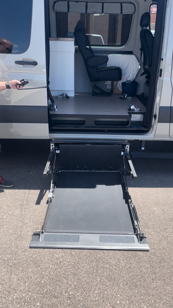 The under-vehicle lift fully deployed and resting flat on the ground beside the van, its platform level with the asphalt and the approach edge down, ready for a chair to roll on.